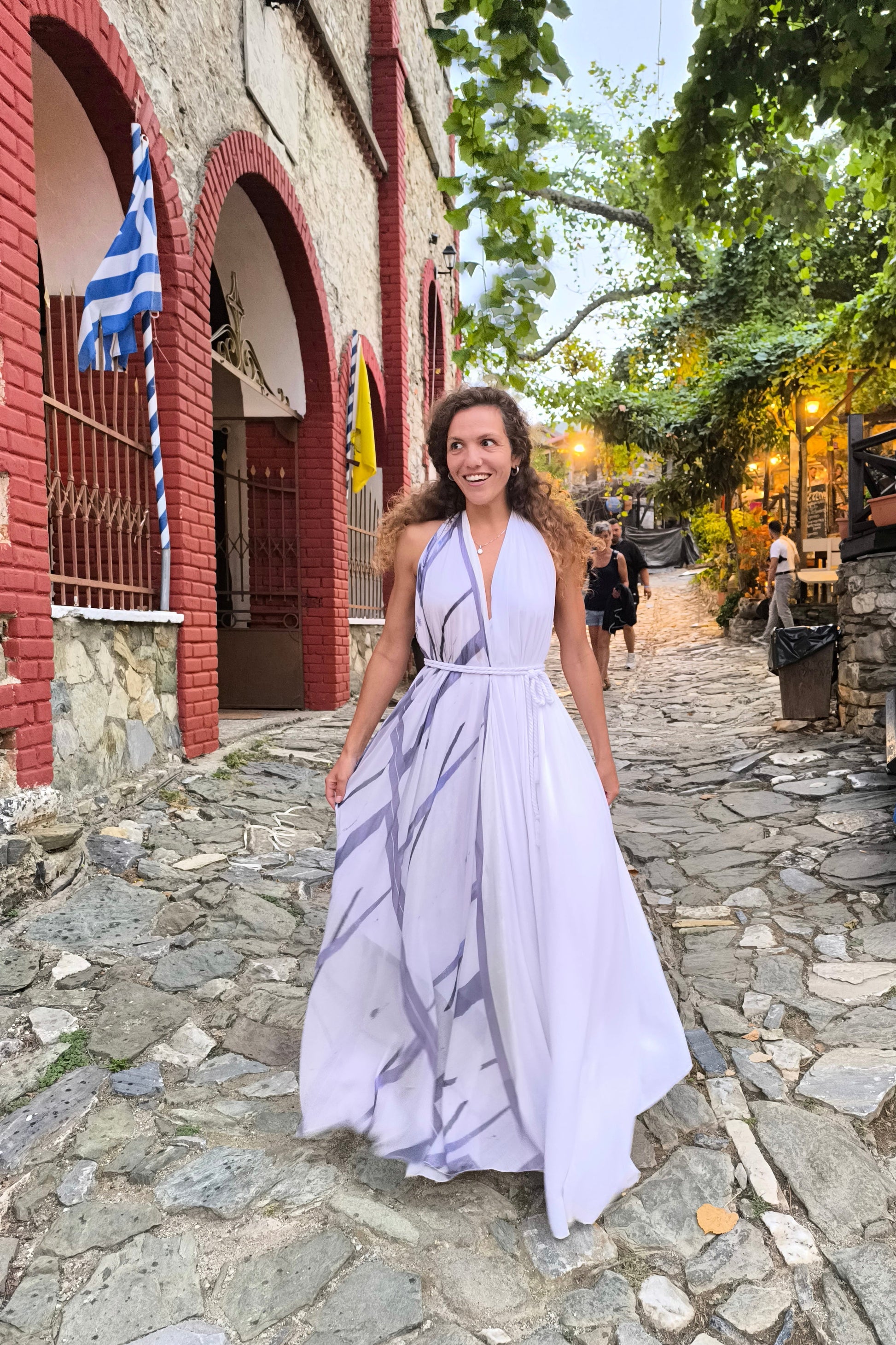 Woman in a purple dress walking on a stone street with a building and trees in the background.
