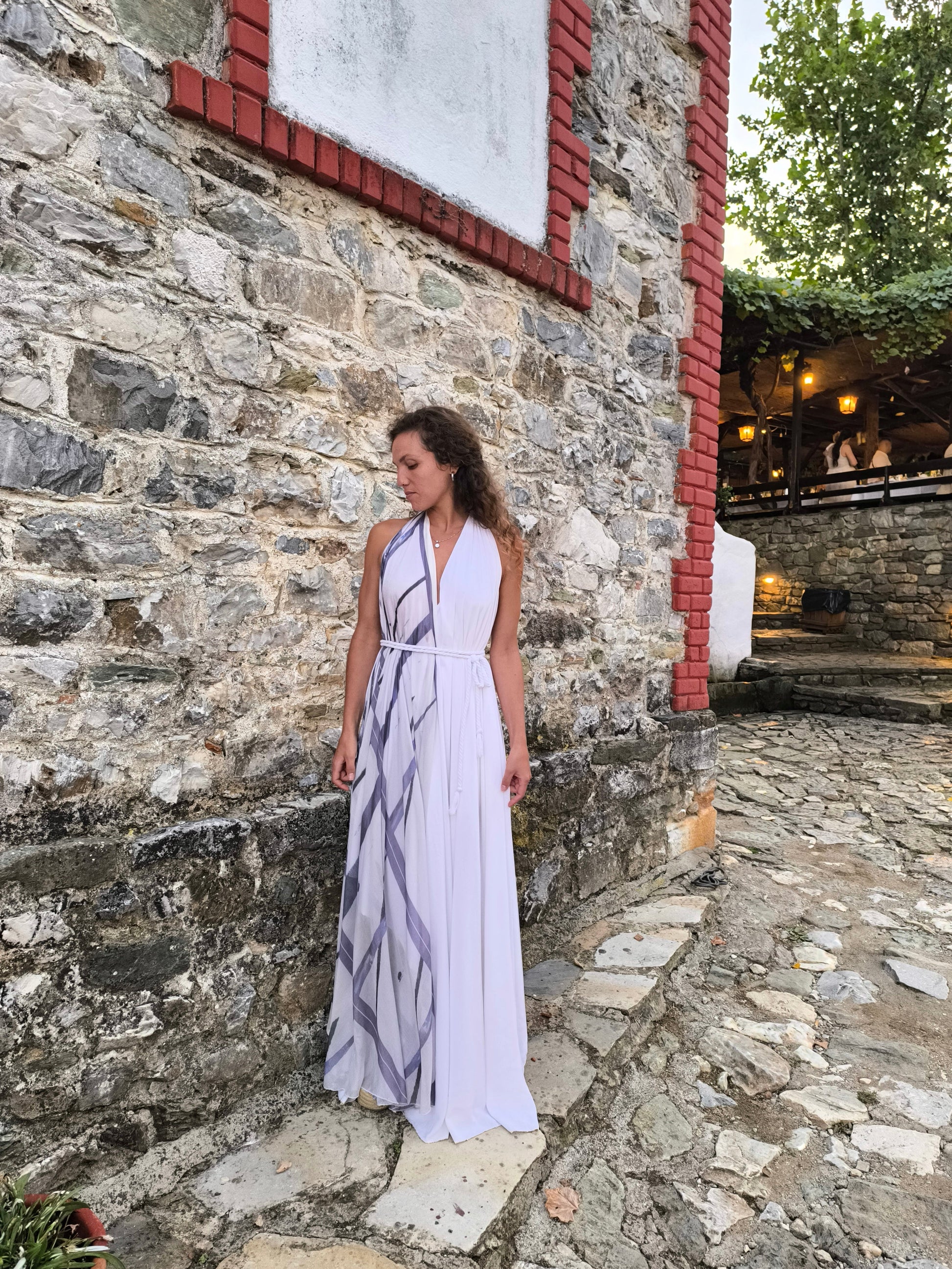 Woman in a white dress standing against a stone wall with a rustic building in the background.